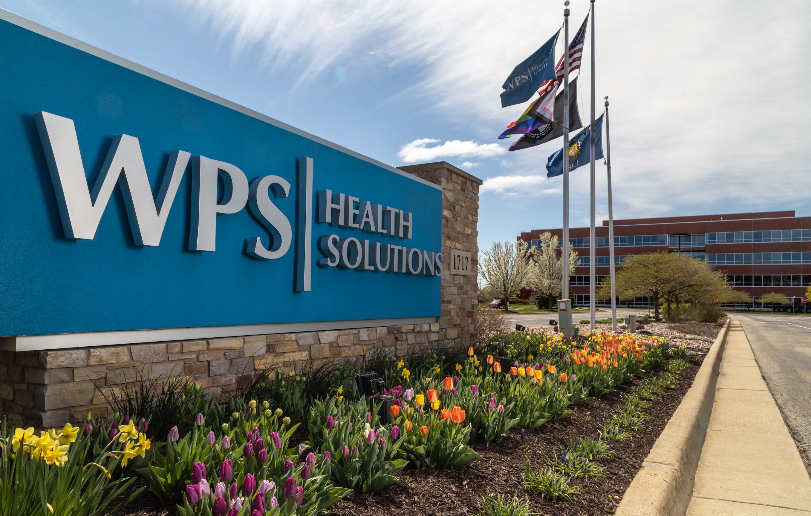 The WPS Health Solutions sign outside of their company headquarters on a bright, sunny day. In the background are numerous flags blowing in the wind and the company headquarters.